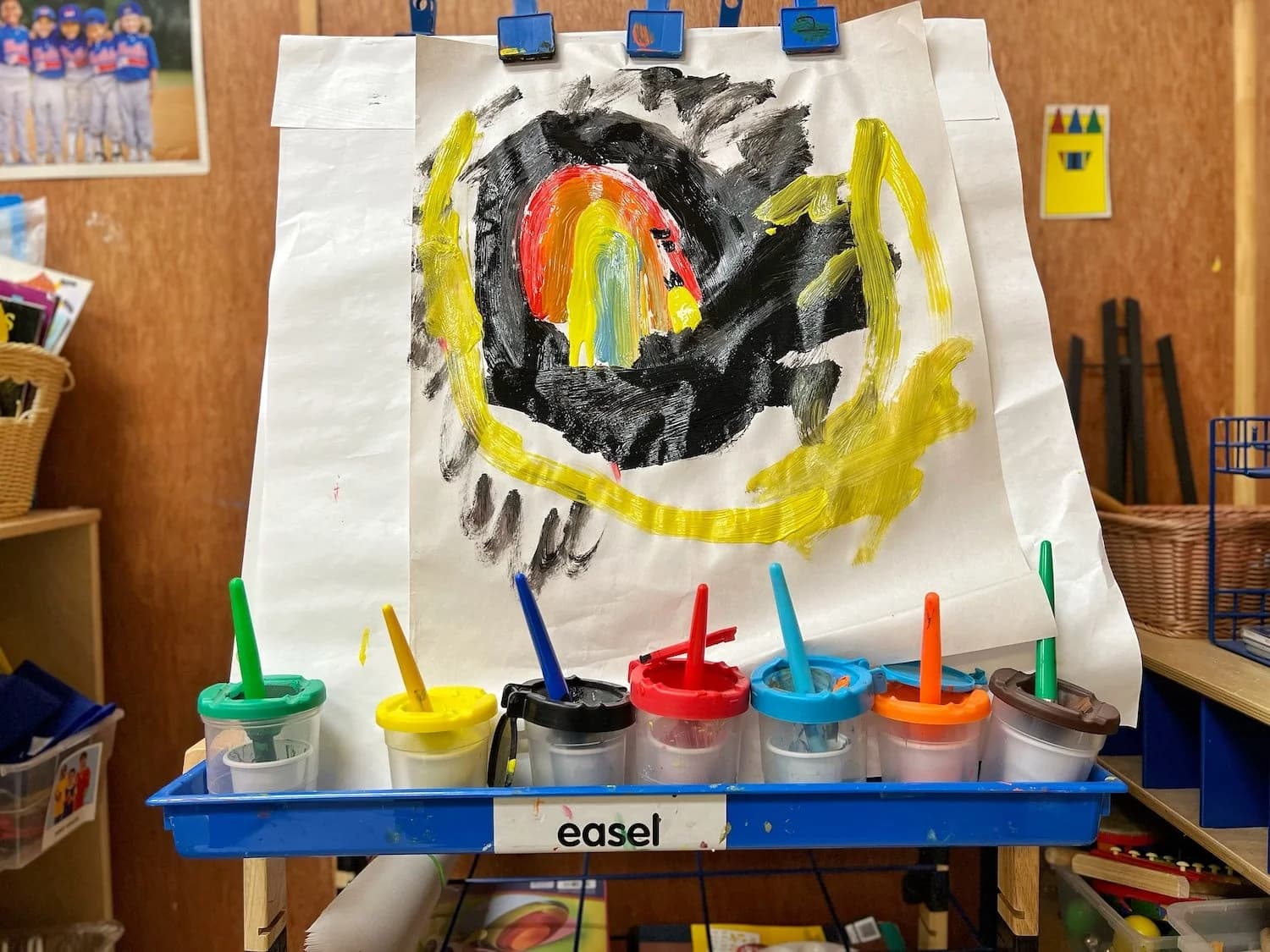 A child's vibrant painting displayed on a classroom easel with paint cups